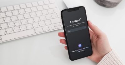 A person holding a smartphone displaying the Qwant search engine homepage, with the tagline "The search engine that values you as a user, not as a product." A white keyboard is visible in the background on a clean, minimalistic desk.